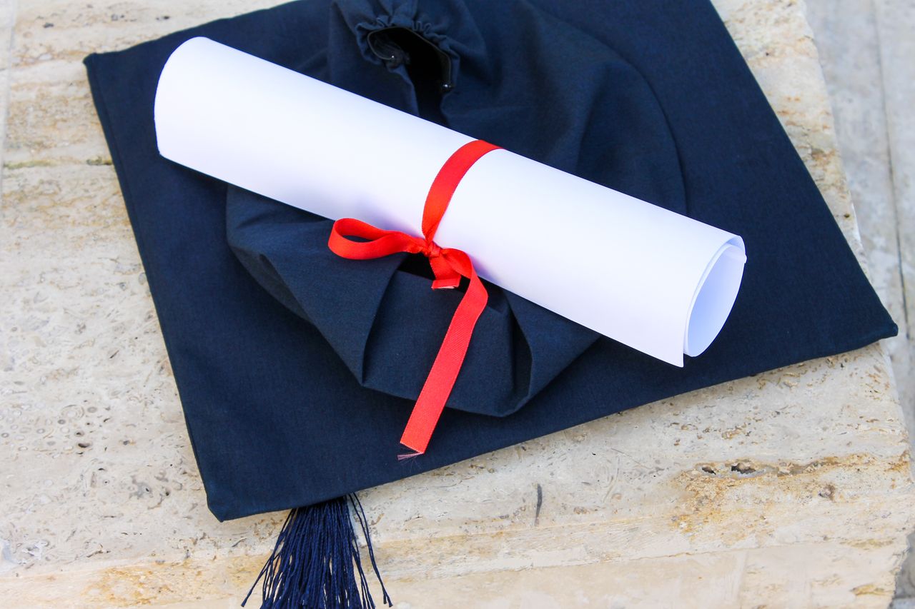 contact-img Graduation cap with diploma roll tied with red ribbon on concrete surface.